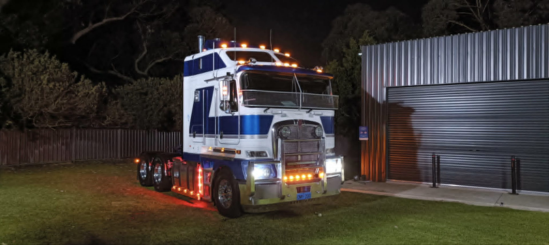 Blue and white cabover prime mover lit up at night beside the depot shed